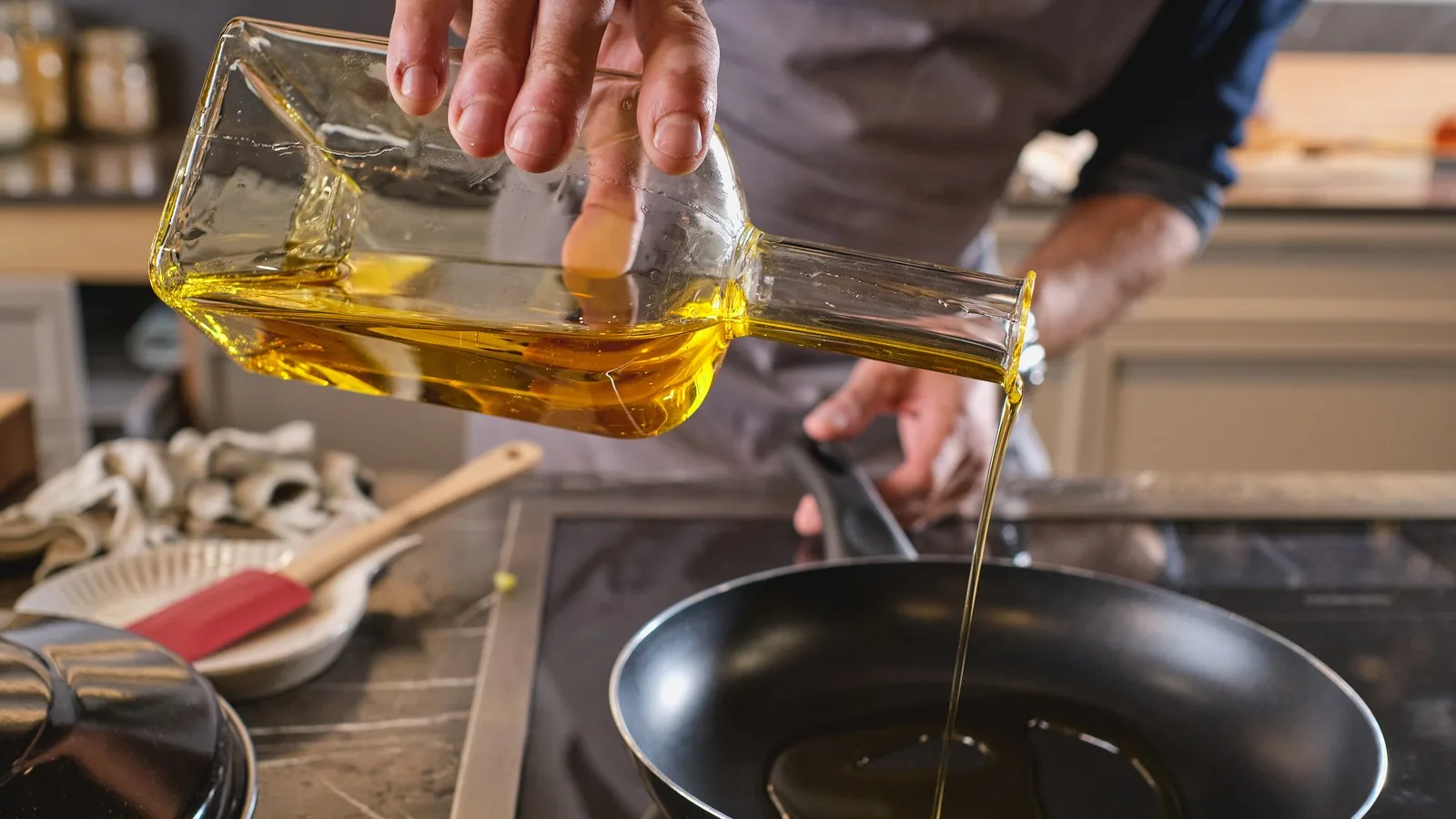 Cooking oil being poured from a glass bottle into a hot frying pan on a stovetop.