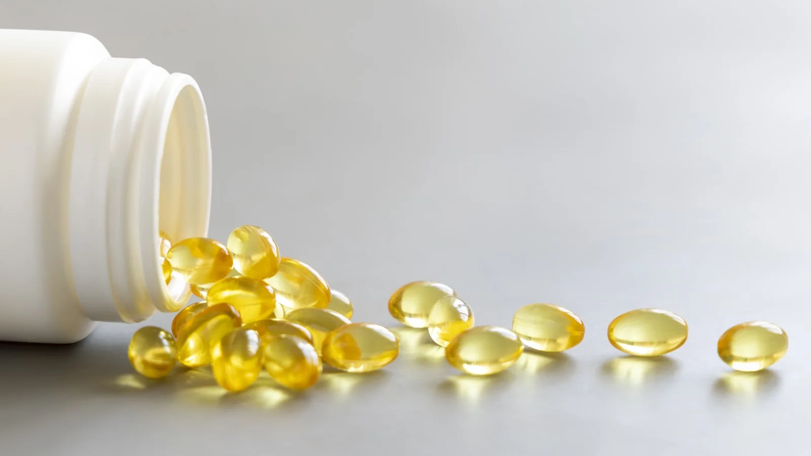 An open supplement bottle on its side with golden softgel capsules spilling out onto a pale grey surface.